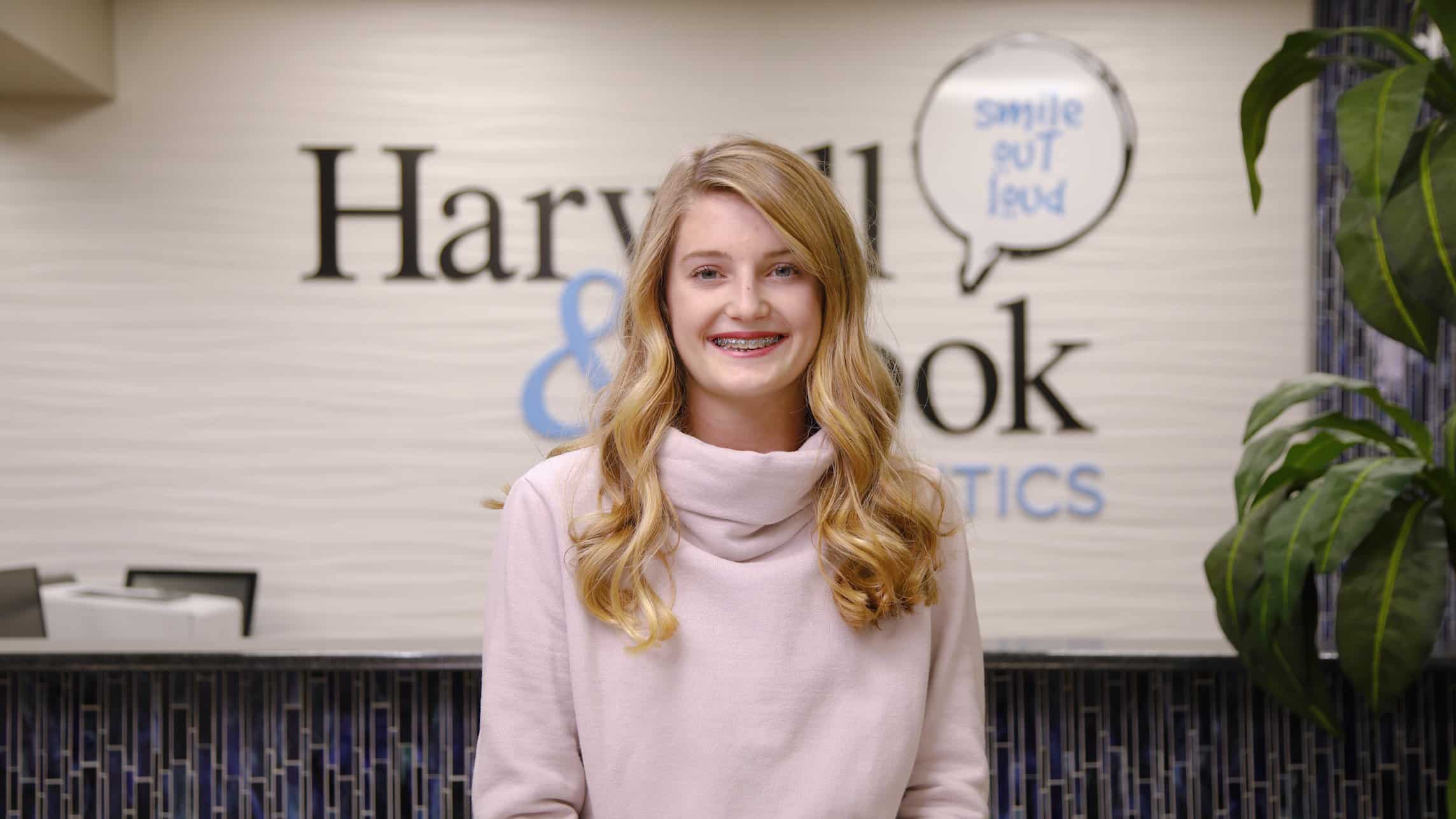 A girl with braces smiling in front of the practice wall. | Harwell & Cook Orthodontics